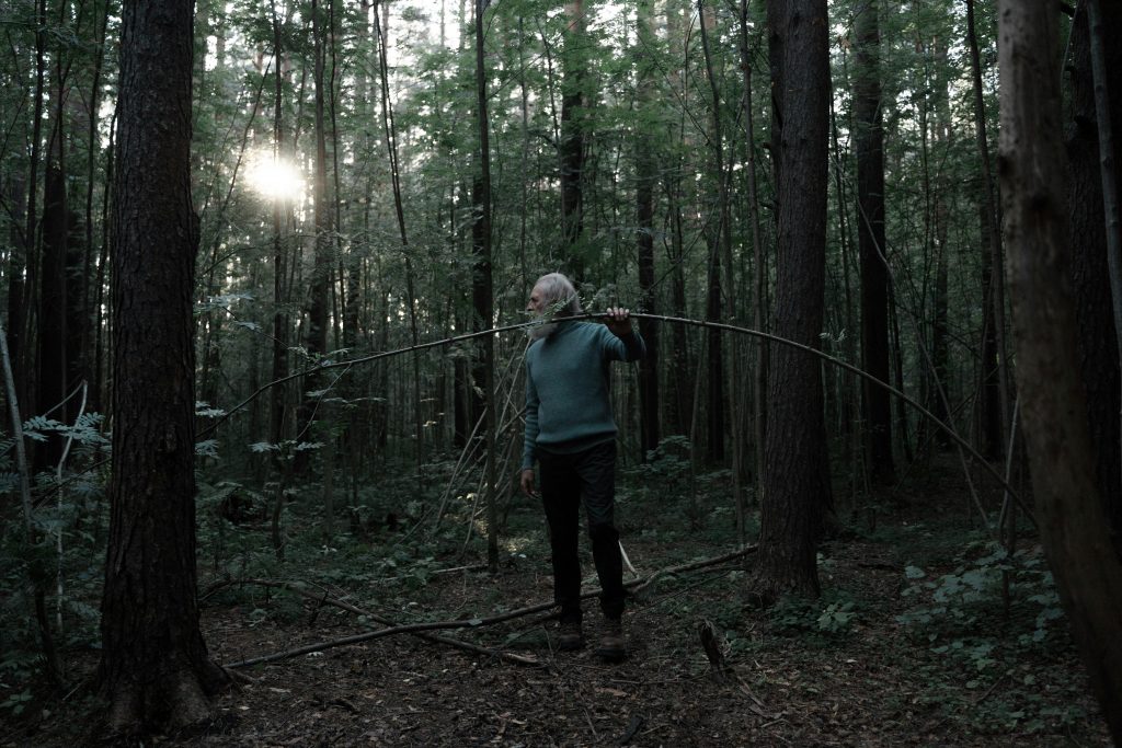 A senior man in a green forest holding a fallen tree branch with sunlight filtering through the trees.